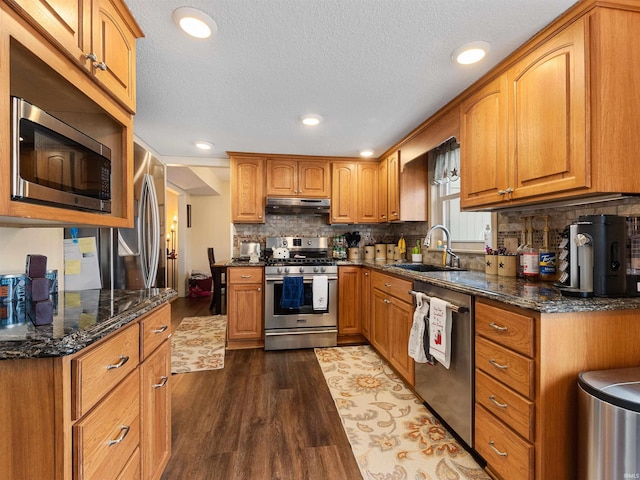 kitchen featuring under cabinet range hood, a sink, dark wood-style floors, appliances with stainless steel finishes, and dark stone counters