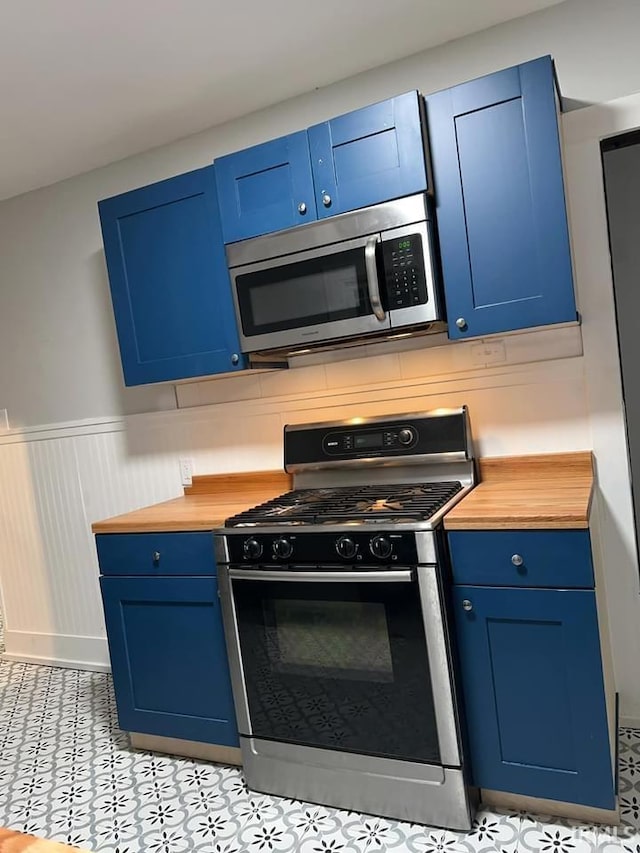 kitchen with butcher block counters, range with gas stovetop, blue cabinetry, wainscoting, and stainless steel microwave