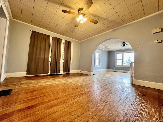 empty room with arched walkways, ornamental molding, a ceiling fan, baseboards, and hardwood / wood-style flooring