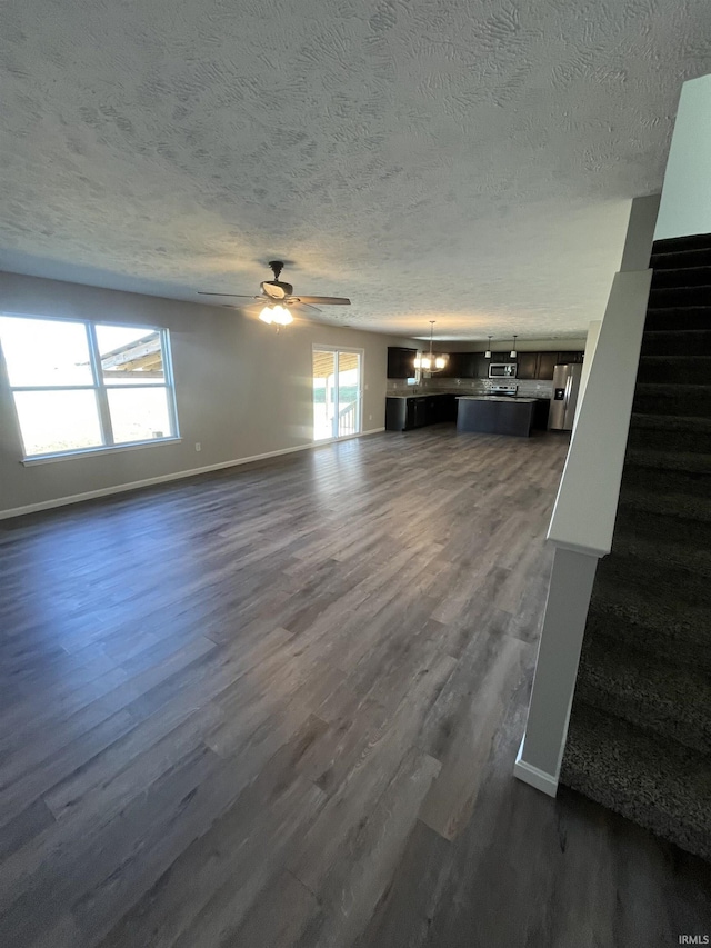 unfurnished living room with dark wood-style floors, baseboards, stairs, a textured ceiling, and ceiling fan with notable chandelier