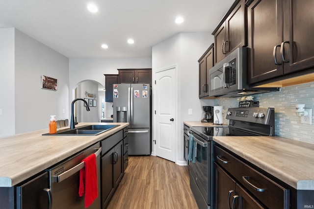 kitchen with wood finished floors, arched walkways, a sink, dark brown cabinets, and appliances with stainless steel finishes