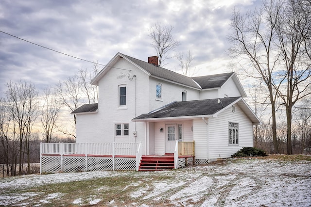 snow covered house with a wooden deck, a chimney, and a shingled roof