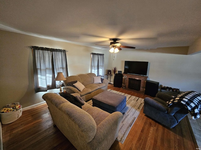 living area featuring a ceiling fan, baseboards, wood-type flooring, a textured ceiling, and a brick fireplace