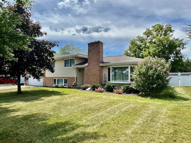 split level home featuring brick siding, fence, roof with shingles, a front yard, and a chimney