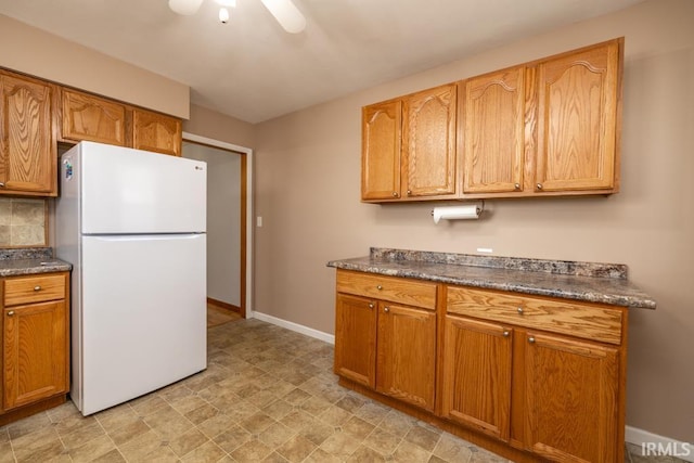 kitchen with backsplash, dark countertops, freestanding refrigerator, brown cabinetry, and baseboards