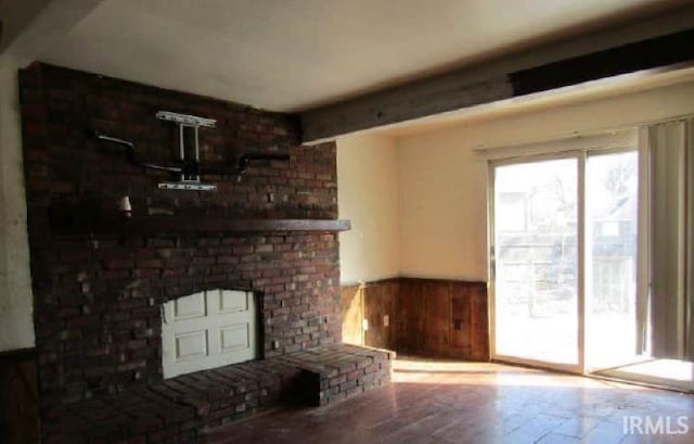 unfurnished living room with beam ceiling, wood finished floors, wood walls, wainscoting, and a brick fireplace