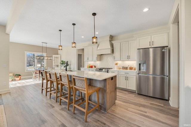 kitchen featuring backsplash, an island with sink, light countertops, custom exhaust hood, and stainless steel appliances