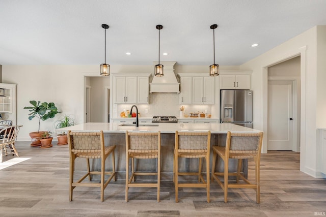 kitchen with a center island with sink, light countertops, custom range hood, range, and stainless steel fridge