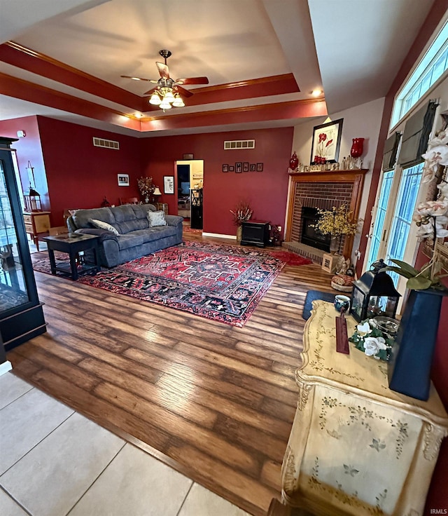 living room featuring visible vents, a brick fireplace, ceiling fan, a tray ceiling, and wood finished floors