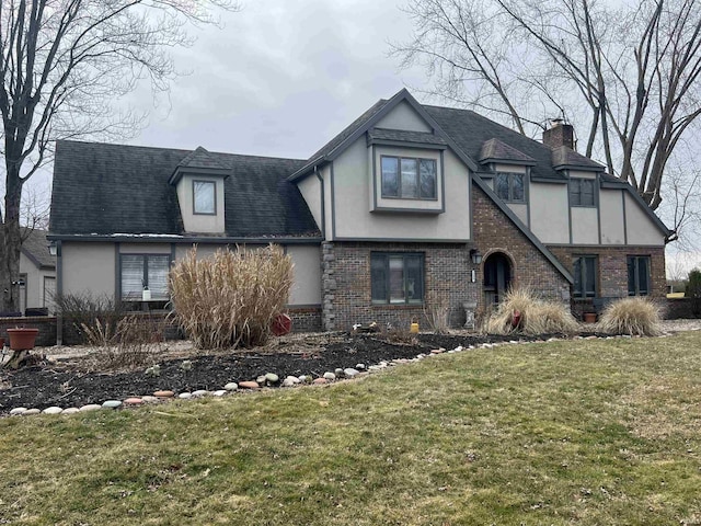 tudor home with stucco siding, brick siding, a front yard, and a shingled roof