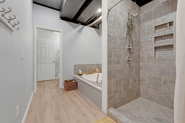 bathroom featuring toilet, wood finished floors, tiled shower, baseboards, and a bath