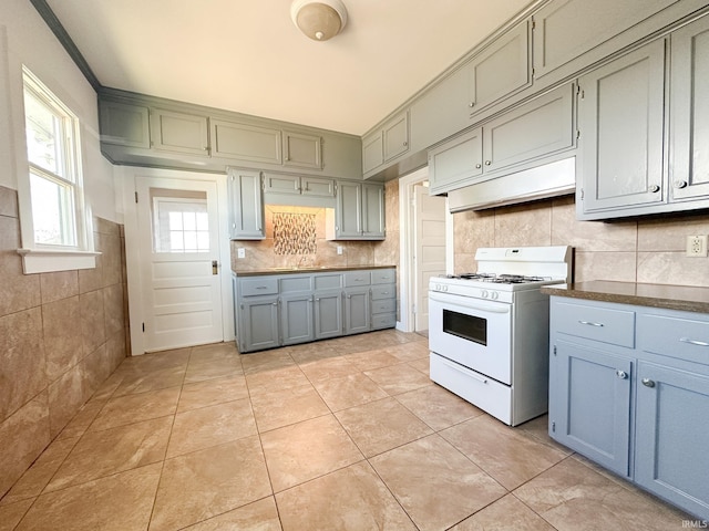 kitchen featuring dark countertops, backsplash, under cabinet range hood, white range with gas stovetop, and light tile patterned flooring