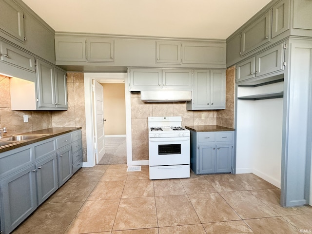 kitchen with white gas stove, under cabinet range hood, dark countertops, light tile patterned flooring, and decorative backsplash