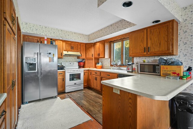 kitchen with wallpapered walls, under cabinet range hood, brown cabinets, appliances with stainless steel finishes, and a peninsula