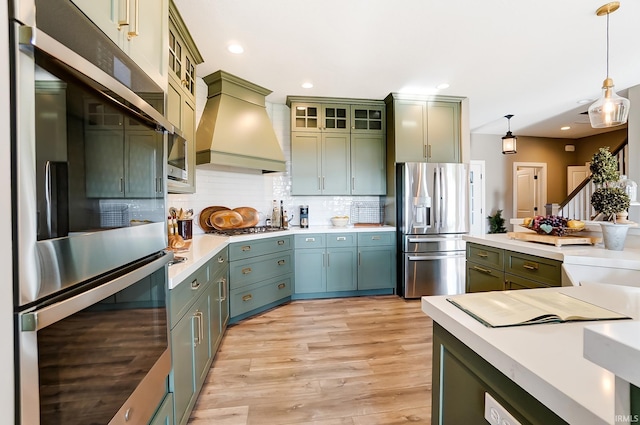 kitchen with light countertops, custom range hood, appliances with stainless steel finishes, green cabinets, and backsplash