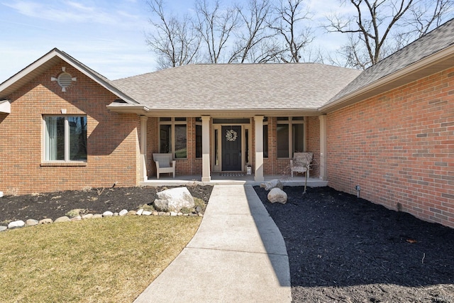 doorway to property with brick siding, covered porch, and roof with shingles