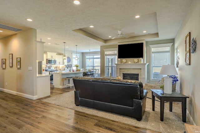 living area with a tray ceiling, a stone fireplace, dark wood-style flooring, and a healthy amount of sunlight