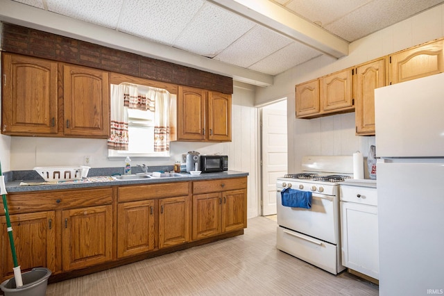 kitchen featuring beam ceiling, a sink, dark countertops, white appliances, and brown cabinetry