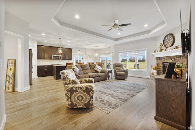 living room featuring baseboards, light wood finished floors, ceiling fan, a stone fireplace, and a raised ceiling