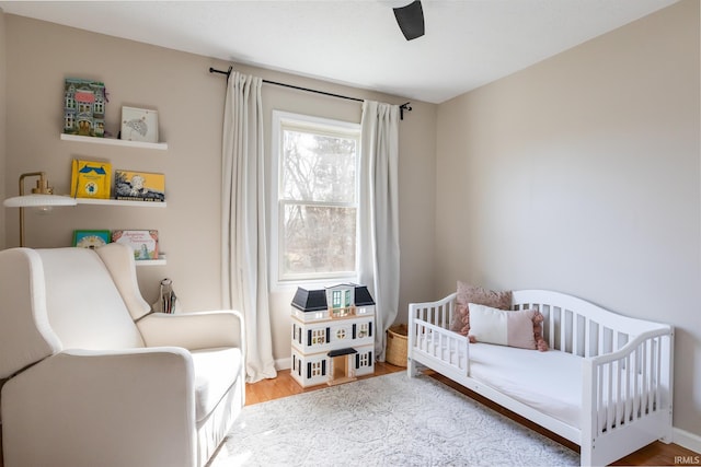 bedroom with baseboards, a ceiling fan, a nursery area, and wood finished floors