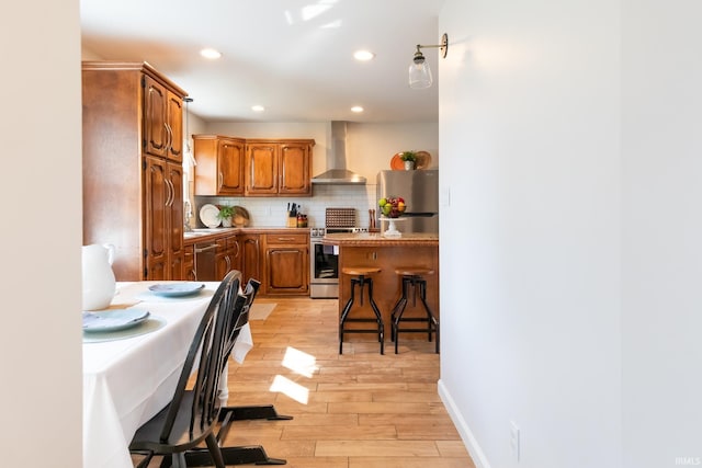 kitchen featuring light wood finished floors, tasteful backsplash, a kitchen bar, stainless steel appliances, and wall chimney exhaust hood