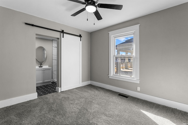 unfurnished bedroom featuring baseboards, ceiling fan, dark carpet, a barn door, and ensuite bath