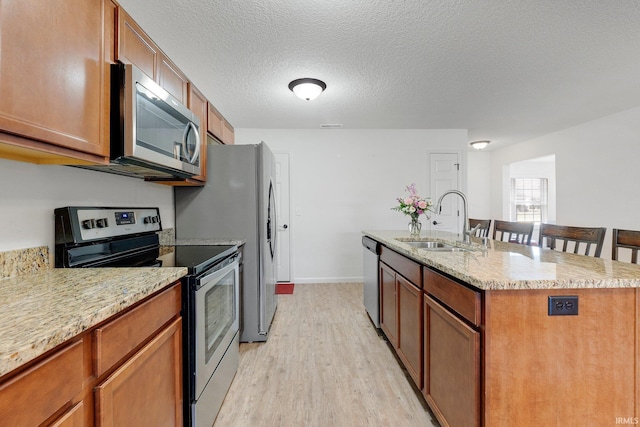 kitchen with light wood-type flooring, an island with sink, light stone counters, stainless steel appliances, and a sink