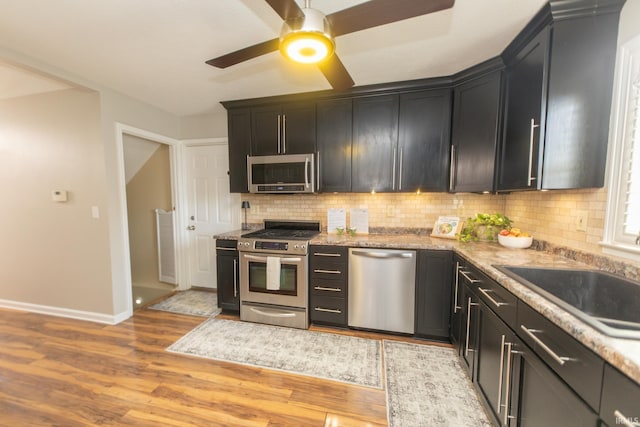 kitchen featuring dark cabinetry, light wood-style flooring, ceiling fan, stainless steel appliances, and tasteful backsplash