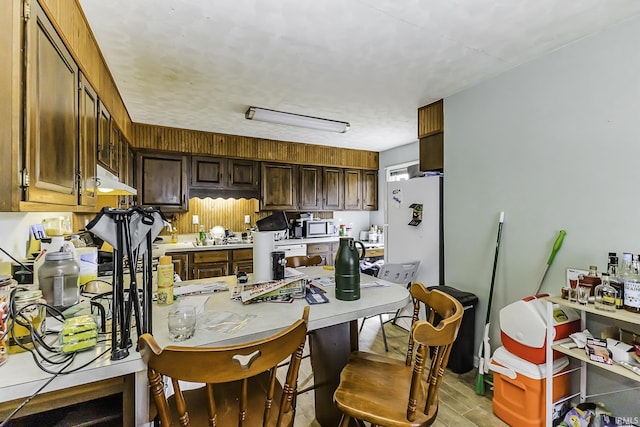 kitchen with under cabinet range hood, stainless steel microwave, freestanding refrigerator, dark brown cabinetry, and light countertops