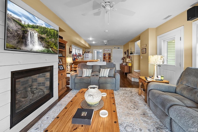 living area with a glass covered fireplace, visible vents, wood finished floors, and ceiling fan