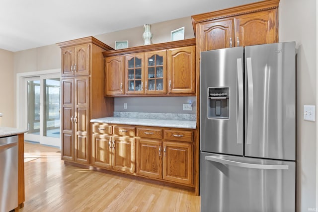 kitchen featuring light wood-type flooring, brown cabinets, stainless steel appliances, glass insert cabinets, and light stone countertops