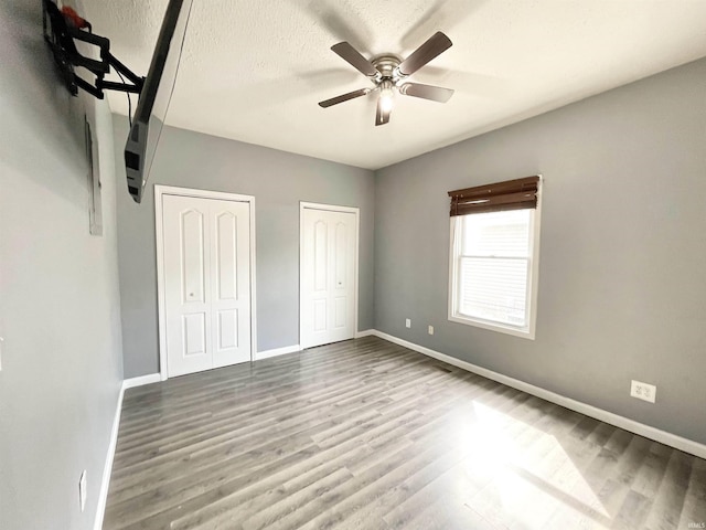unfurnished bedroom featuring two closets, a ceiling fan, a textured ceiling, wood finished floors, and baseboards