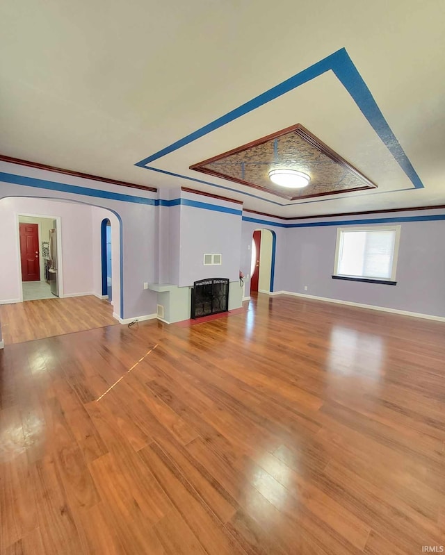 unfurnished living room with a tray ceiling, wood finished floors, arched walkways, and ornamental molding