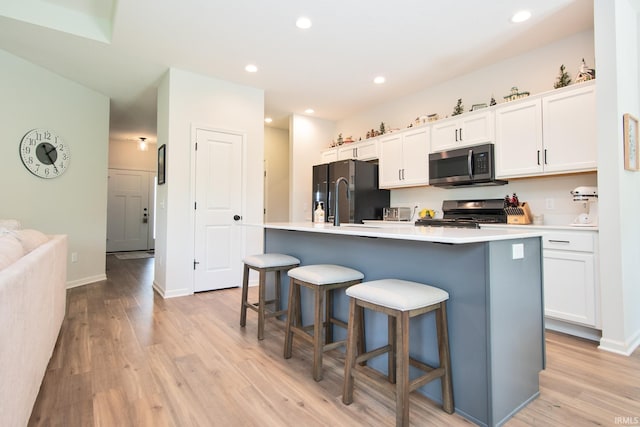 kitchen featuring an island with sink, stove, stainless steel microwave, light wood-type flooring, and black refrigerator with ice dispenser