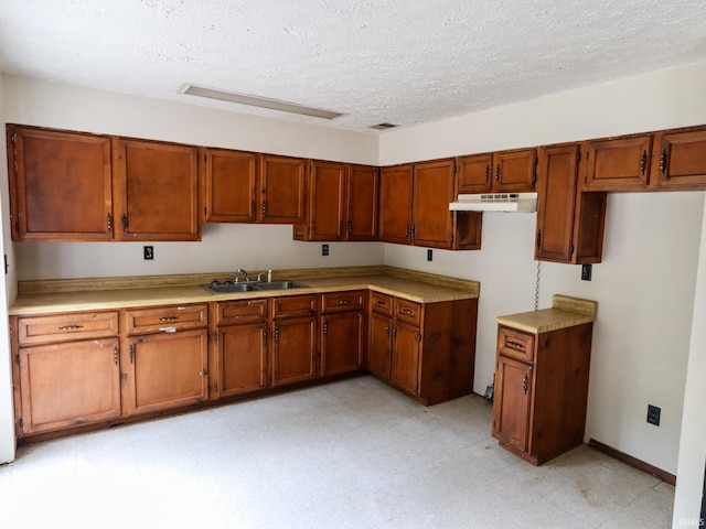 kitchen featuring under cabinet range hood, light floors, brown cabinets, a textured ceiling, and a sink