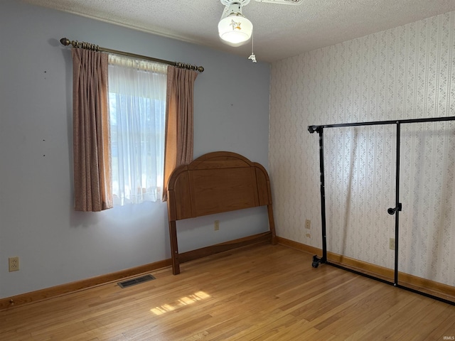 unfurnished bedroom featuring a textured ceiling, visible vents, light wood-style flooring, baseboards, and wallpapered walls