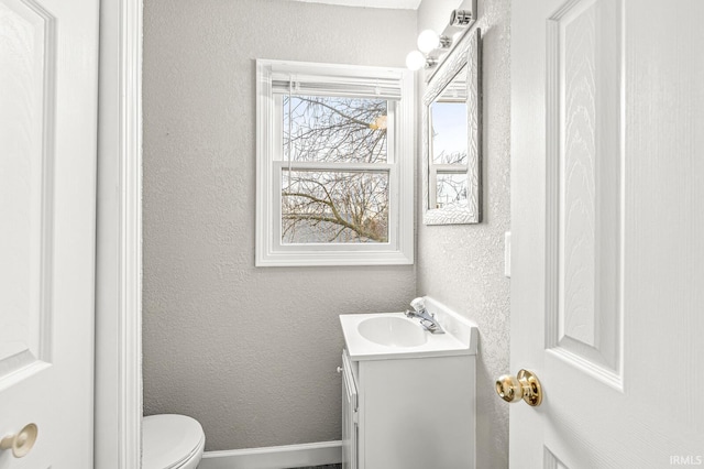half bath featuring vanity, toilet, a textured wall, and baseboards