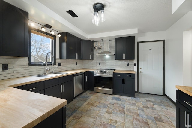 kitchen with decorative backsplash, wall chimney exhaust hood, butcher block counters, appliances with stainless steel finishes, and a sink