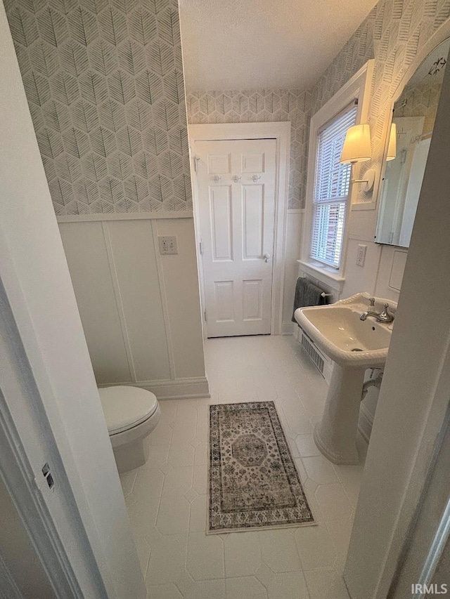 bathroom with tile patterned flooring, toilet, wallpapered walls, a decorative wall, and wainscoting