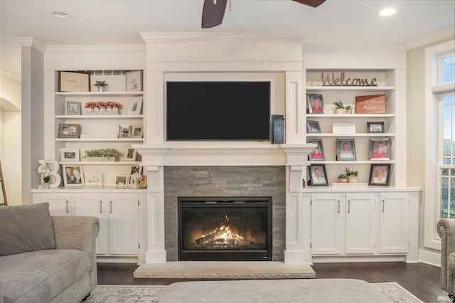 living room featuring dark wood-style flooring, crown molding, a ceiling fan, built in features, and a glass covered fireplace