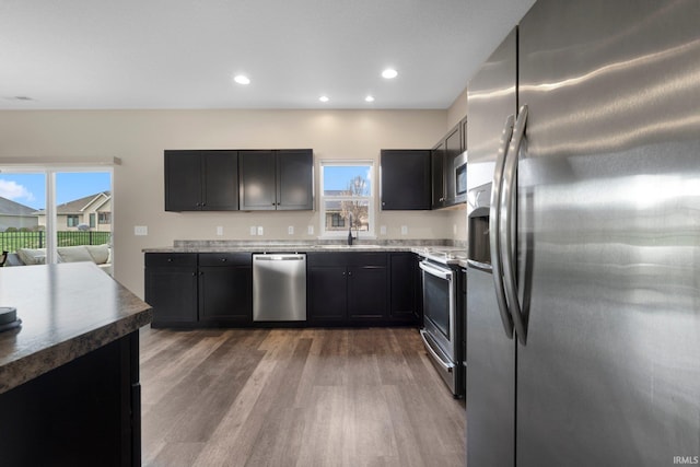 kitchen with appliances with stainless steel finishes, light wood-style floors, dark cabinets, recessed lighting, and stone countertops
