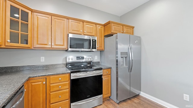 kitchen with dark countertops, appliances with stainless steel finishes, glass insert cabinets, and light wood-style flooring