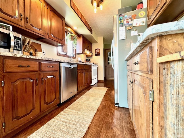 kitchen featuring a textured ceiling, dark wood-style flooring, brown cabinetry, under cabinet range hood, and appliances with stainless steel finishes