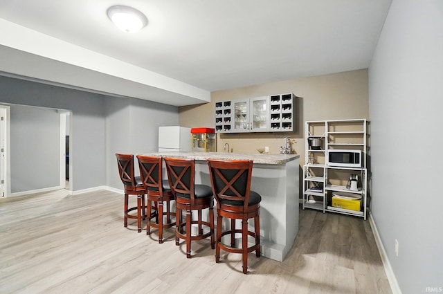indoor wet bar with white microwave, light wood-style flooring, and baseboards