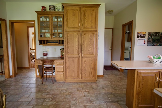 kitchen featuring glass insert cabinets, light countertops, brown cabinets, stone finish flooring, and baseboards