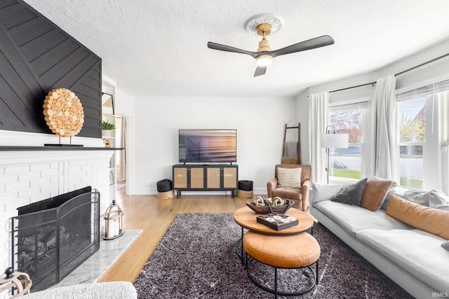 living room with wood finished floors, a brick fireplace, baseboards, a ceiling fan, and a textured ceiling