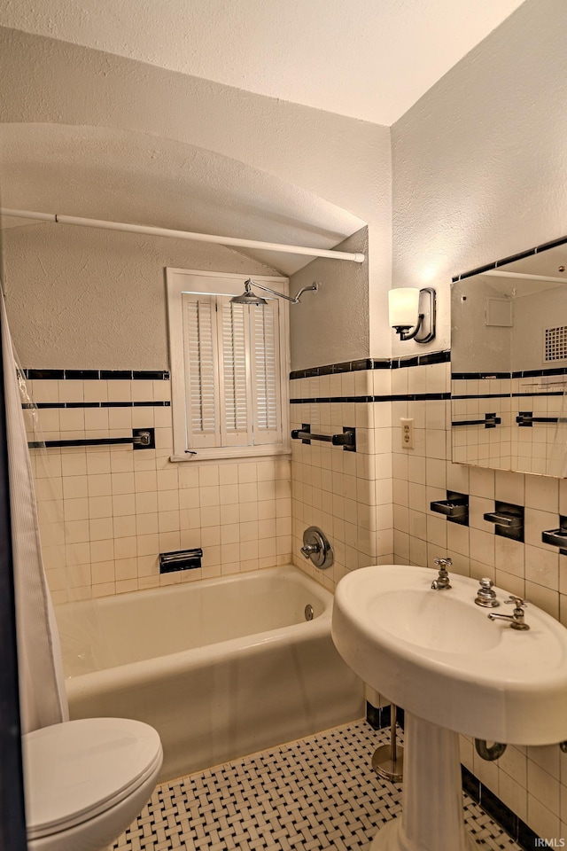 full bath featuring shower / bath combo with shower curtain, a textured wall, toilet, tile walls, and a wainscoted wall