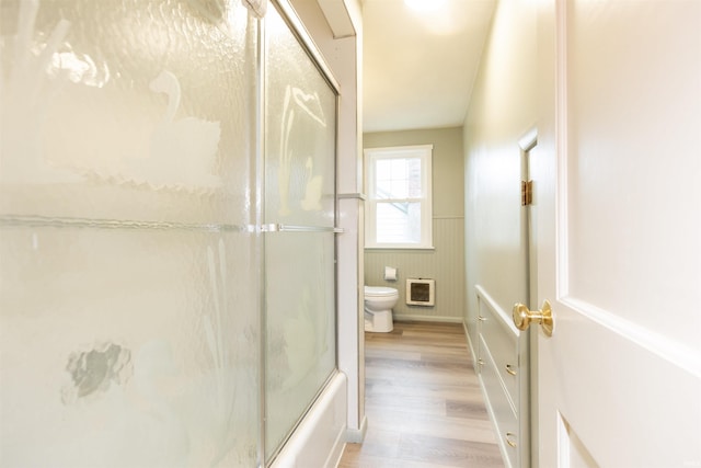 bathroom with shower / bath combination with glass door, wood finished floors, toilet, and a wainscoted wall
