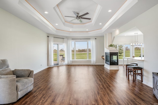 living room with a multi sided fireplace, baseboards, ceiling fan with notable chandelier, dark wood-style floors, and a tray ceiling