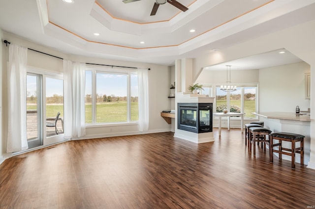 living area with crown molding, dark wood-style floors, a tray ceiling, a multi sided fireplace, and ceiling fan with notable chandelier